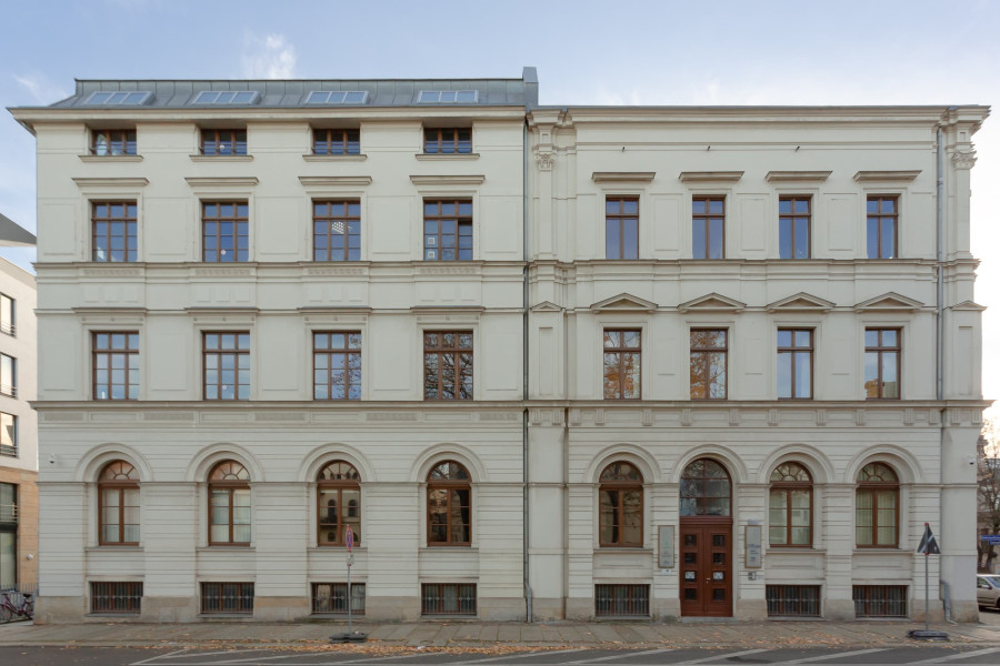 Ein Eckgebäude mit einer Fassade aus der Gründerzeit; mit hohen runden Festern im Erdgeschoss, zwei Stockwerken und einem Dachgeschoss; die Eingangstüre befindet sich nicht mittig, sondern im rechten Bereich des Gebäudeabschnitts.