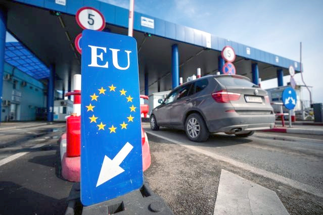 Das Foto zeigt einen Grenzübergang zwischen Bulgarien und Rumänien, im Vordergrund eine blaue Tafel mit der Beschriftung "EU" und zwölf im Kreis angeordneten gelben Sternen und einem großen Richtungspfeil