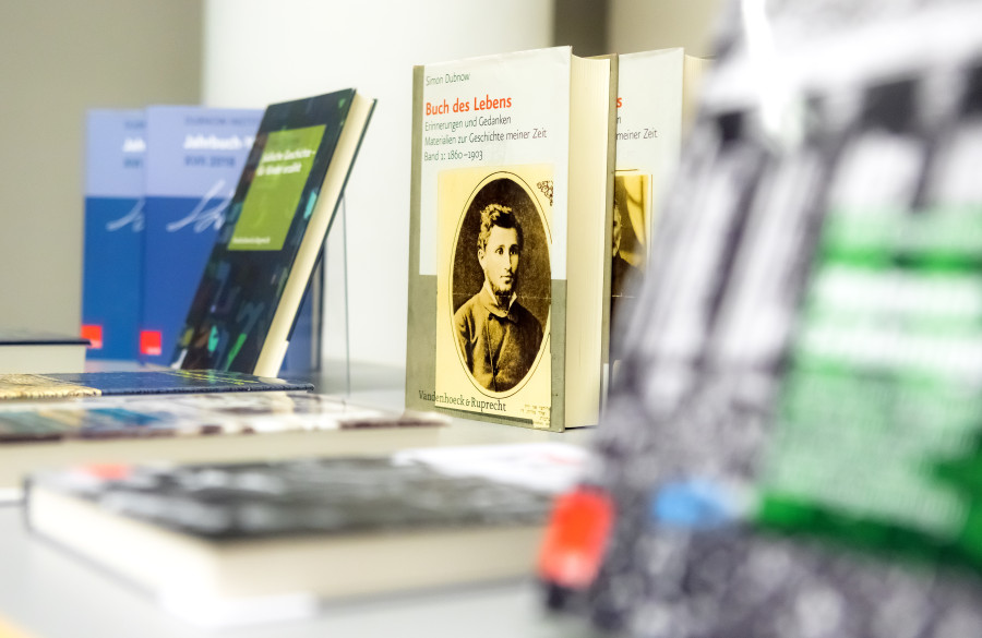 Ein Tisch, auf dem die Publikationen des Dubnow-Instituts präsentiert werden; einige Buchcover lassen sich nur schemenhaft erkennen; zentral in der Mittel des Bildes ist der erste Band des "Buch des Lebens" von Simon Dubnow zu sehen.