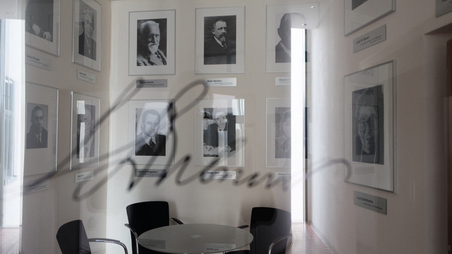 Durch eine Glasscheibe fotografiert ist ein Durchgangsraum in einem Bürogebäude zu erkennen mit einem runden Tisch und drei Stühlen außenherum; an der Wand hängen Schwarz-Weiß-Fotografien mit Porträts berühmter jüdischer Gelehter.